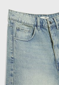 Gros plan sur un jean en denim bleu clair montrant la poche avant, les passants de ceinture, le bouton et la fermeture éclair, avec des coutures et une texture de tissu visibles.