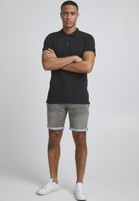 Juodas polo marškinėliai, pilkos džinsinės šortai, sukritusios rankovės. Balti sportiniai bateliai. Paprastas dizainas, be matomų raštų. Stovinti poza su sukryžiuotomis rankomis.