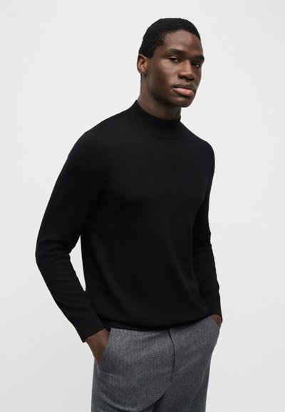 Νεαρός άνδρας με τα χέρια στις τσέπες, φορώντας μαύρο πουλόβερ με μακριά μανίκια και ζιβάγκο, καθώς και γκρι παντελόνι, κοιτάζει προς την κάμερα.