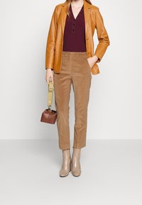 Vrouw in een camel leren blazer, bordeauxkleurige knopenboven, tan corduroy broek, beige enkellaarsjes, met een kleine bruine handtas met een beige schouderband.