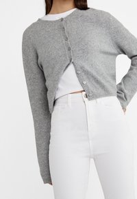 Cardigan gris en maille avec un col rond, doté d'une fermeture à boutons sur le devant, manches longues et un design court, associé à un jean blanc.