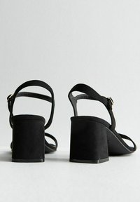 Sandalias de tacón de ante negro con tacón bloque, puntera abierta y correas finas para soporte en el tobillo. Diseño minimalista con una textura suave.