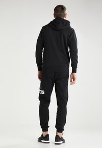 Uomo che indossa un hoodie nero e pantaloni sportivi neri Adidas, in piedi con le spalle rivolte verso una sfondo chiaro e uniforme.