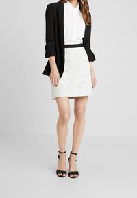 Blazer noir, chemise blanche à boutons, jupe mini blanche texturée et escarpins noirs à lanières ; look superposé avec des couleurs et des matériaux contrastants.