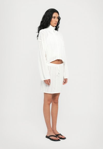 White knit top with a mock collar and cropped design, paired with a matching drawstring skirt. Both feature intricate patterns. Black flip-flops.