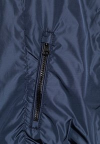 Dunkelblauer Pufferjackenbereich mit einem vertikalen schwarzen Reißverschluss, glatter Textur, mit Falten im Bereich des Reißverschlusses und einer gesteppten Verarbeitung.