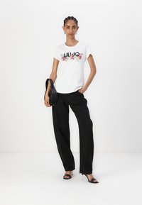 T-shirt en coton blanc avec un graphique floral portant l'inscription "LIU JO," associé à un pantalon large noir et des sandales à talons noires. Sac à bandoulière noir.