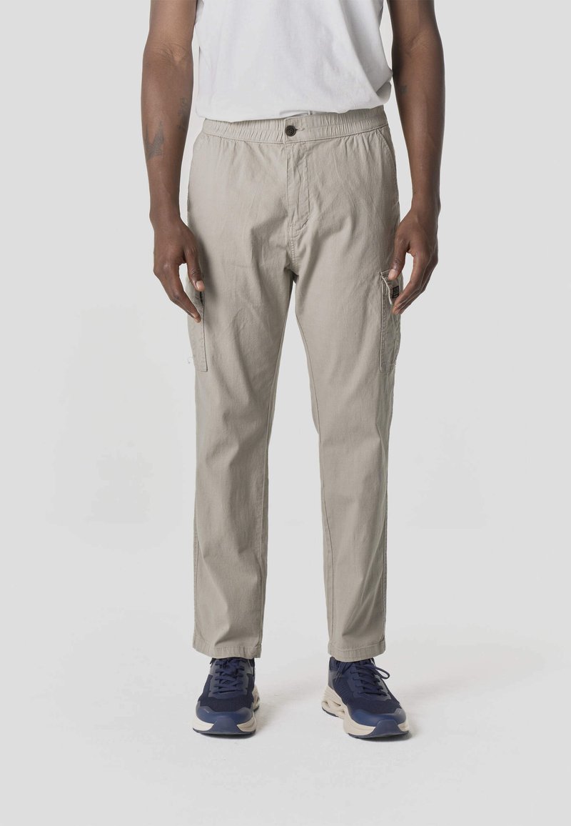 Hombre con pantalones cargo beige, camiseta blanca y zapatillas azul marino, de pie contra un fondo liso de color gris claro.