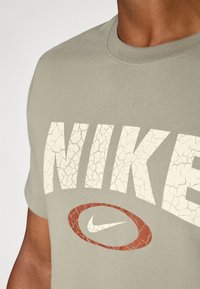 Olivenfarget Nike T-skjorte med store, sprukne kremfargede bokstaver og et slitt rødt logoaktsent. Den har rund hals og korte ermer.
