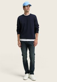 Suéter de cuello redondo navy, gorra azul claro, jeans de mezclilla oscura y zapatillas blancas. Conjunto casual con un ajuste holgado y diseño minimalista.