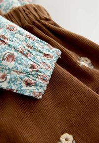 Een bruine corduroy stof met een gestructureerd oppervlak, met bovenaan een bloemenpatroon in lichtblauw en bruin.