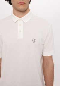 Hvid polo-shirt lavet af struktureret stof, med en klassisk krave, tre knapper og et lille broderet logo på venstre bryst.