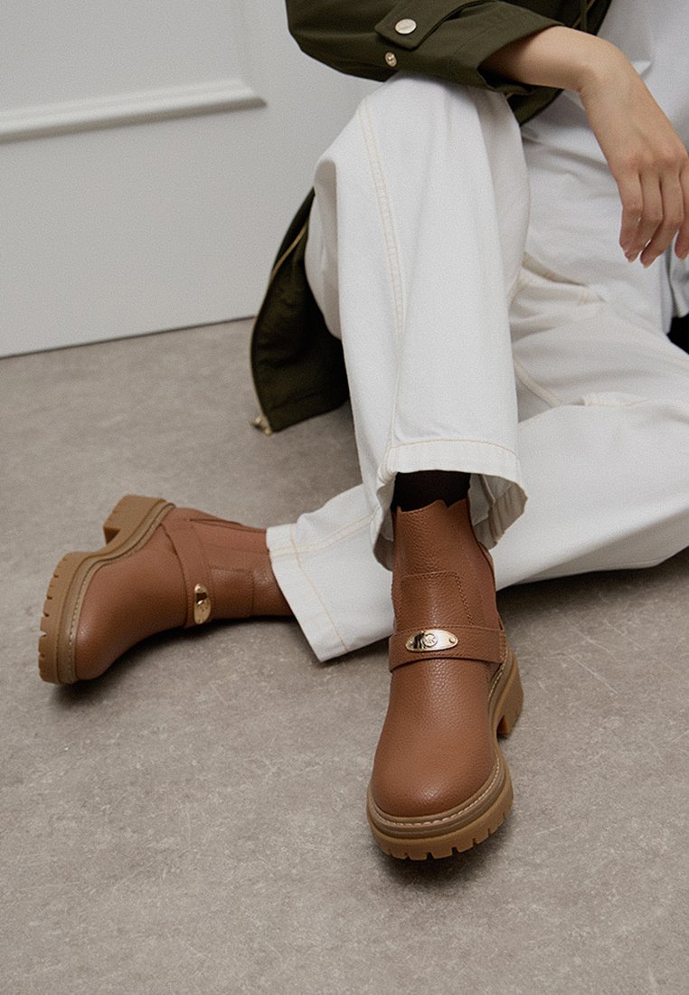 Bruine enkellaarzen van textuur synthetisch materiaal, met een gouden gespaccent en een chunky rubberen zool. G gedragen met witte broeken.