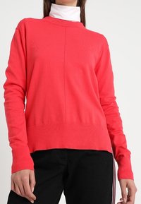 Pull en corail à manches longues, coupe droite, ourlet côtelé et col rond, superposé à un col roulé blanc.