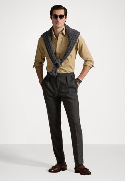 Cămașă bej cu nasturi, pulover gri din tricot cu model, așezat pe gât, pantaloni gri croiți, și pantofi din piele maro. Ochelari de soare negri purtați.