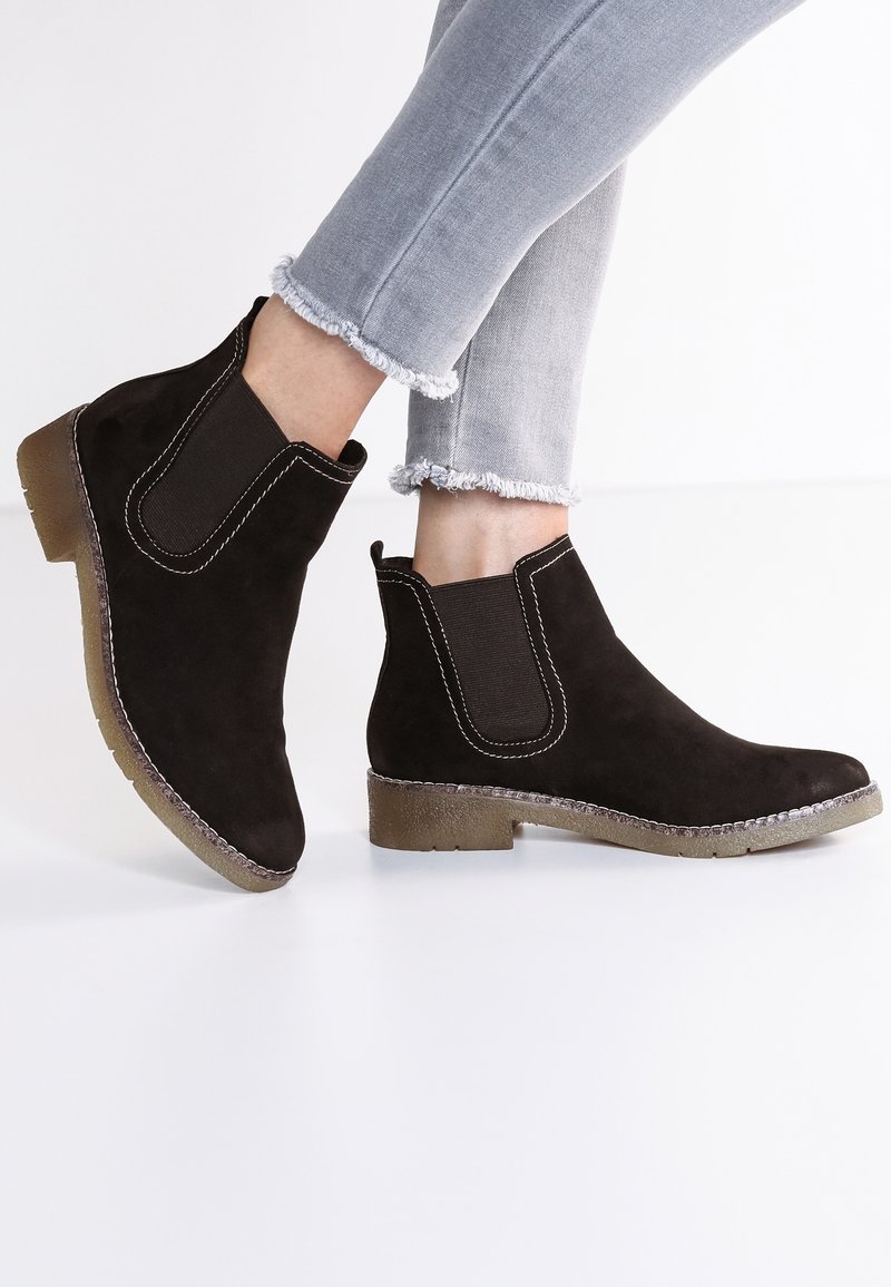 Bottines noires en daim à talons bas avec panneaux élastiques latéraux portées avec un jean gris clair à ourlet effiloché sur fond blanc.