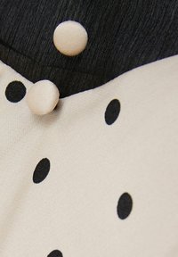 Beige tyg med stora svarta prickar, med en svart övre del och två beige tygklädda knappar. Slät textur, platt design.