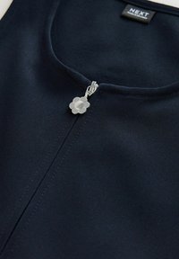 Mornarska zip-up oblačila z okroglo izrezom, s srebrnim zadrgo v obliki cvetlice in teksturirano tkanino. Oznaka prikazana na vrhu.