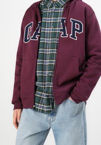 Sudadera con cremallera color granate con letras blancas "GAP", usada sobre una camisa de cuadros verde y azul, combinada con vaqueros azul claro. Tejido de mezcla de algodón.