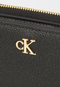 Portefeuille en cuir noir texturé avec une fermeture éclair dorée et un logo "CK" doré en relief sur le devant. Une vue rapprochée met en évidence le matériau et les détails.