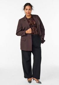 Blazer marron sur un top bordeaux foncé avec des motifs floraux, associé à un pantalon large noir et des escarpins argentés. Matériaux texturés.