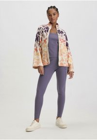Fleece vest met ritssluiting in oranje, paars en beige met abstract patroon, gecombineerd met een lila tanktop en leggings, afgewerkt met witte sneakers.