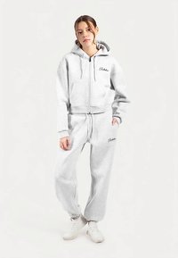 Heathergrijze cropped hoodie met een ritssluiting aan de voorkant, grote zakken en logo; bijpassende sweatpants met elastische manchetten en logodetails; gedragen met witte sneakers.