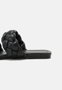 Sandales mules en cuir noir avec une sangle tressée, semelle plate et texture lisse, adaptées à une tenue décontractée.