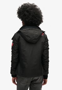Chaqueta negra aislante con capucha, bolsillos con cremallera y acentos de logo rojo en los hombros. Fabricada con un material suave y texturizado.