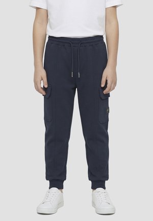 Personne portant un pantalon cargo jogging bleu marine avec des poignets et une taille élastiques, accompagné de baskets blanches et d'une chemise blanche à manches courtes.