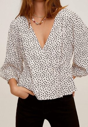 Blouse portefeuille blanche à pois noirs irréguliers, manches ballon trois-quarts, décolleté en V profond, associée à un pantalon noir et des colliers de perles superposés.