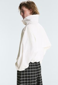 Jeune personne aux cheveux courts portant une veste blanche à col montant et un pantalon à carreaux noir et blanc, vue de profil, tournée vers la gauche.