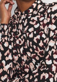 Blusa con cuello en un tejido negro con un estampado de leopardo en tonos de rosa, blanco y burdeos. Presenta detalles fruncidos en los puños.