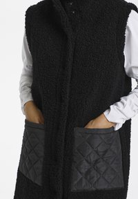 Gilet nero realizzato in tessuto texturizzato con cappuccio, dotato di due tasche frontali imbottite a diamante e bottoni sul davanti.