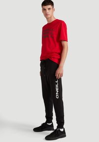 Rød T-shirt med sort trykt logo, parret med sorte sweatpants med hvid "O'Neill" tekst. Modellen bærer sorte sneakers.