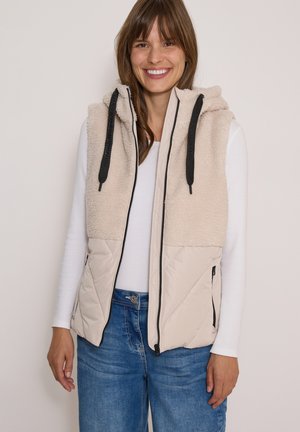 Donna sorridente che indossa un gilet in pile beige con cappuccio sopra una camicia bianca a maniche lunghe e jeans blu, in piedi davanti a uno sfondo semplice.