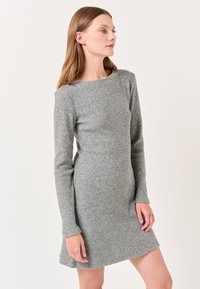 Femme aux cheveux châtain clair portant une robe tricotée ajustée à manches longues et arrivant au genou, se tenant devant un fond neutre.