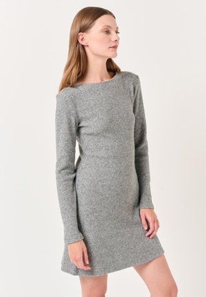 Frau mit hellbraunem Haar, die ein langärmliges, knielanges, eng anliegendes graues Strickkleid trägt, steht vor einem neutralen Hintergrund.