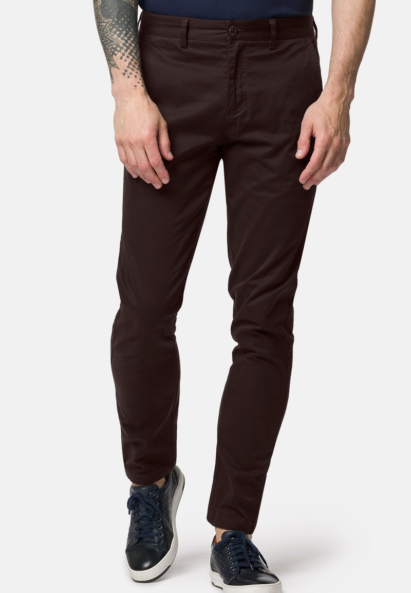 Ciemnobrązowe, slim-fit spodnie wykonane z mieszanki bawełny, z płaskim przodem, bocznymi kieszeniami i gładką teksturą. Łączone z granatowymi trampkami.