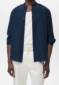 Merenkulkija napitettava paita, jossa on mandariinikaulus, valmistettu pehmeästä kankaasta. Paita on päälle puettu valkoisen pyöreäkaula-aukkopaidan yli ja yhdistetty vaaleansävyisiin housuihin.