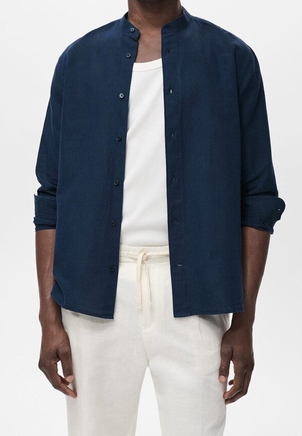 Merenkulkija napitettava paita, jossa on mandariinikaulus, valmistettu pehmeästä kankaasta. Paita on päälle puettu valkoisen pyöreäkaula-aukkopaidan yli ja yhdistetty vaaleansävyisiin housuihin.