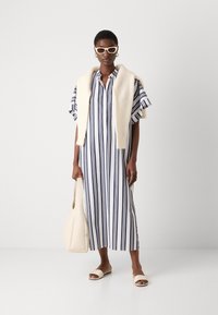 Abito maxi a righe in bianco e blu navy, con maniche corte e bottoni sul davanti, abbinato a una sciarpa crema e una borsa beige. Tessuto liscio e fluido.