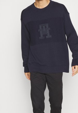 Mann trägt einen marineblauen Strickpullover mit einem hervorgehobenen doppelten "TH"-Logo auf der Brust und schwarze Hose vor hellem, einfarbigem Hintergrund.