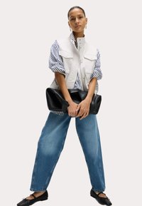 Gilet sans manches blanc sur une chemise rayée bleue et blanche, associé à un jean large bleu clair et des chaussures plates noires, tenant un sac noir.