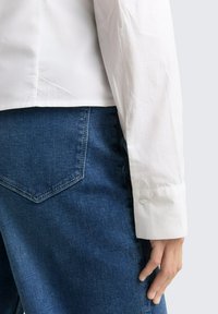 Wit overhemd met lange mouwen en een gladde textuur, gecombineerd met donkerblauwe denim jeans met een diamantvormige achterzakdetail.