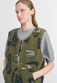 Zeleni flis prsluk s plavim Nike logotipima i teksturiranim detaljima, s patentnim zatvaračem sprijeda, dva džepa, nošen preko sive majice.
