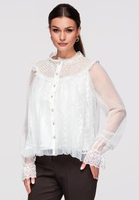 Femme portant un chemisier en dentelle blanche transparente avec des boutons en perle, un col montant et de longues manches bouffantes, associé à un pantalon foncé.
