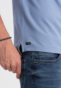 Světle modré polo tričko s texturovanou tkaninou, bočními ventilačními otvory a malou černou značkovou nášivkou. Nosené s tmavými denimovými džíny.