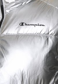 Silver puffjacka med en glansig yta, med en svart dragkedja och "Champion"-logotypen tydligt visad i svart text.