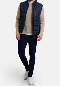 Marineblaue gepolsterte Weste mit vertikalen Nähten und einer Reißverschlusstasche, getragen über einem hellbeigen Pullover. Kombiniert mit dunklen Jeans und weißen Sneakern.